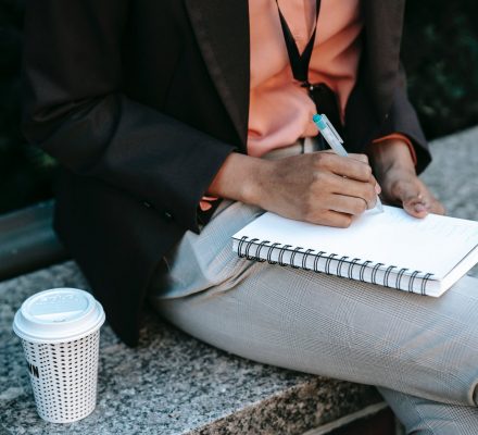 ethnic woman writing notes in planner with takeaway coffee
